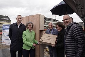 Gruppe von Menschen bei einer Grundsteinlegung mit Holzbox und Schild, im Hintergrund ein Bauplatz.