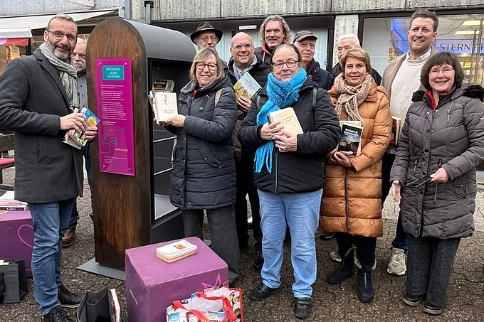 Eine Gruppe von Menschen steht vor dem Bücherschrank im Maubishof und hält Bücher in den Händen, lächelnd und fröhlich.