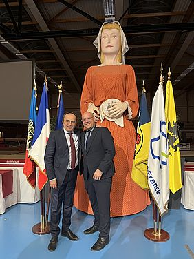 Zwei Männer (Sébastien Leprêtre und Christian Horn-Heinemann) stehen neben einer großen Statue einer Frau in einem orangefarbenen Kleid.