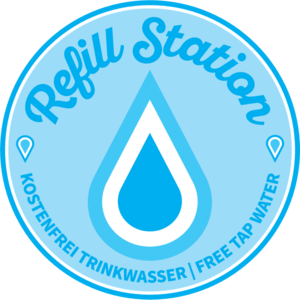 Ein blaues Rundzeichen mit der Aufschrift "Refill Station" und "Kostenfrei Trinkwasser / Free Tap Water" mit einem Wassertropfen in der Mitte.