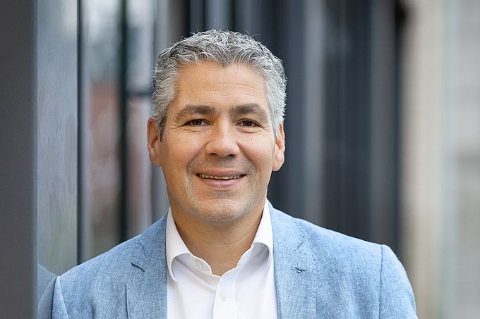 Ein lächelnder Mann mit grauem Haar trägt ein hellgraues Sakko, ein weißes Hemd und eine braune Gürtelschnalle.