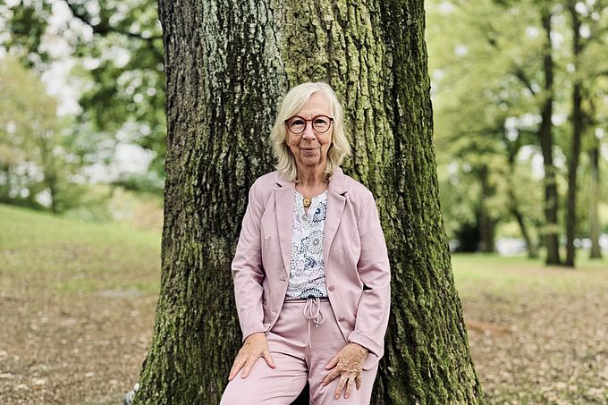 Eine Frau sitzt lässig an einem Baum in einem Park, gekleidet in einem pinken Anzug und weißen Sneakern.