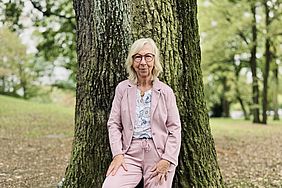 Eine Frau sitzt lässig an einem Baum in einem Park, gekleidet in einem pinken Anzug und weißen Sneakern.