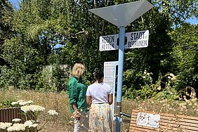 Zwei Personen lesen ein Informationsschild in einem begrünten Stadtgebiet mit Wildblumen und Schildern auf Deutsch.