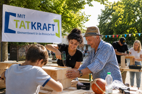 Eine Gruppe von Menschen arbeitet gemeinsam an einem DIY-Projekt im Freien, unterstützt durch das "MIT TATKRAFT"-Programm.