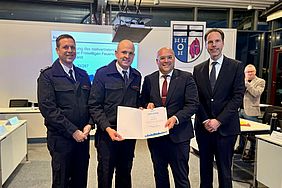 Vier Männer in Anzügen und Uniformen halten ein Zertifikat und lächeln für ein Gruppenfoto.