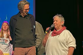 Drei Personen auf einer Bühne, eine ältere Frau spricht in ein Mikrofon, daneben ein Mann und eine junge Frau.