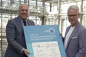 Zwei Männer halten ein blaues Dokument mit Unterschriften, das "Pakt für Planungsbeschleunigung im Rheinischen Revier" betitelt ist.