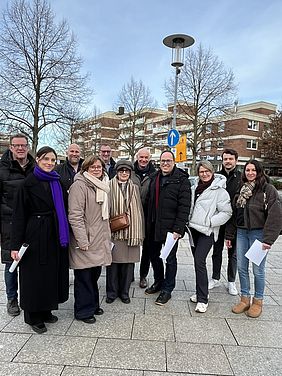 Eine Gruppe von Menschen steht draußen und lächelt in die Kamera, vor einem Gebäude und leeren Bäumen.