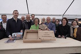 Gruppe von Menschen bei einer Grundsteinlegungszeremonie mit einem Holzbehälter und einer Plakette.