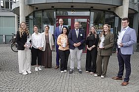 Gruppe von acht Personen vor einem Gebäude, die lächelnd posieren und kleine Geschenke halten.