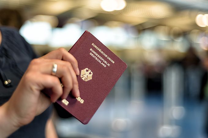 Eine Hand hält einen deutschen Reisepass vor einem unscharfen Hintergrund.