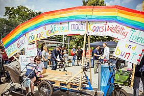 Eine bunte Regenbogenstruktur mit deutschen Slogans auf einem belebten Marktplatz mit Menschen und Fahrrädern.