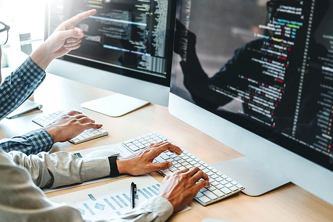Zwei Personen arbeiten an Computern mit Datenvisualisierungen auf den Bildschirmen, wobei eine Person auf etwas zeigt.
