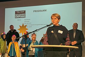 Eine Frau spricht bei einer Veranstaltung mit dem Titel "Neujahrsempfang" vor einem Publikum, hinter ihr stehen Kinder in Kostümen.