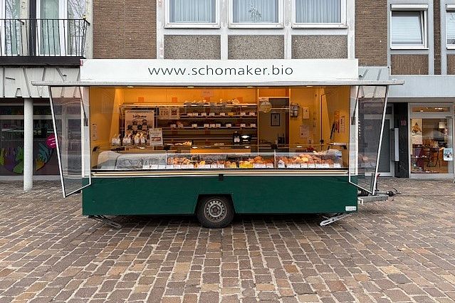 Ein mobiler Imbisswagen mit der Website "www.schomaker.bio" auf einem Kopfsteinpflasterplatz vor einem Gebäude.