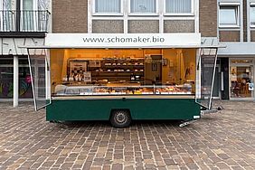 Ein mobiler Imbisswagen mit der Website "www.schomaker.bio" auf einem Kopfsteinpflasterplatz vor einem Gebäude.