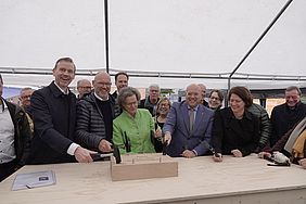 Eine Gruppe von Menschen bei einer Grundsteinlegungszeremonie mit Schaufeln und einem Holzblock.