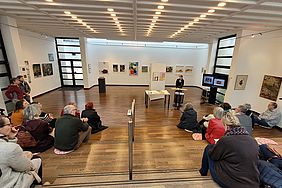 Eine Kunstausstellung mit einer Gruppe von Menschen, die einer Präsentation zuhören. Bilder hängen an den Wänden.