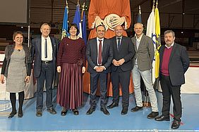Gruppe von sieben Personen (Violett Massiet Zielinski, Rainer Wilking, Tanja Klein-Kohlstette, die beiden Bürgermeister Sébastien Leprêtre und Christian Horn-Heinemann, der Kulturausschussvorsitzende Ulrich Orlinski, Foudil Tarzoult) steht vor einer großen Statue in einem Innenraum mit Flaggen im Hintergrund.