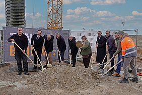 Gruppe von Menschen bei einem symbolischen Spatenstich auf einer Baustelle mit Schaufeln und einem Kran im Hintergrund.