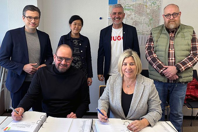 Auf dem Bild zu sehen: Daniel Mang (ALHO Systembau GmbH) und Bürgermeisterin Ursula Baum zeichnen die Verträge im Beisein von Sebastian Trautermann (ALHO), Wing-Yee Leung, Michael Wilms und Bryan McDonough (alle Stadt Kaarst) 
