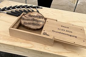 Ein Holzkasten mit Werkzeugen und einer beschrifteten Holzscheibe, die ein Datum und einen Namen trägt, auf einer Baustelle.