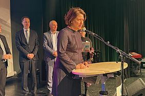 Landrätin Katharina Reinhold spricht an einem Podium mit Mikrofon, während mehrere Personen im Hintergrund stehen.